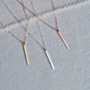 May include: Three delicate gold necklaces with thin chains and vertical bar pendants. The pendants are different lengths and have varying finishes, including smooth, textured, and hammered.