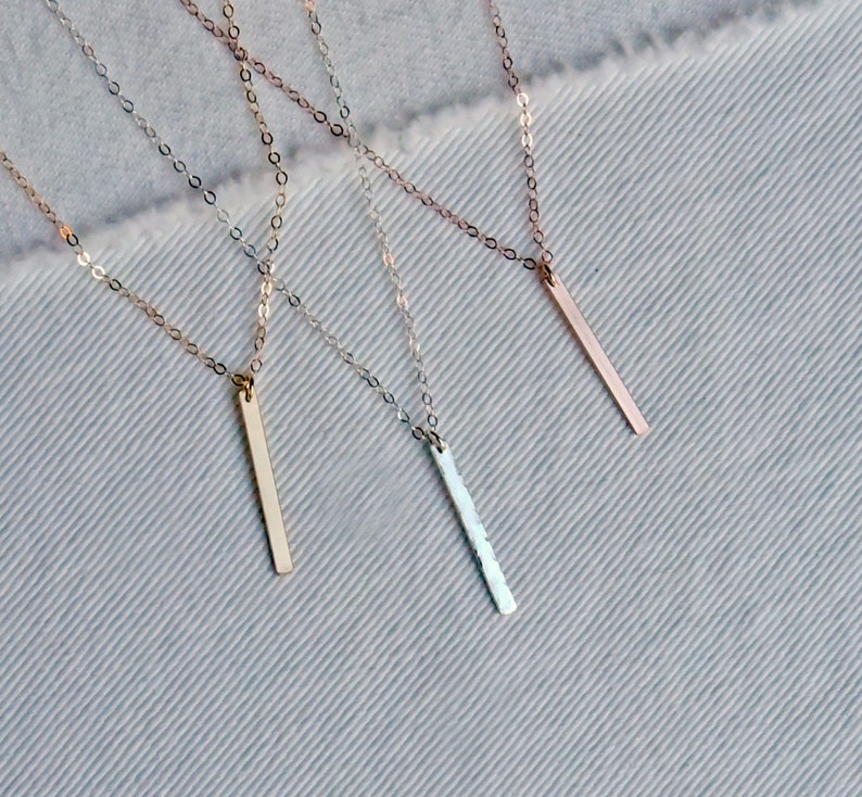 May include: Three delicate gold necklaces with thin chains and vertical bar pendants. The pendants are different lengths and have varying finishes, including smooth, textured, and hammered.