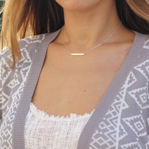 May include: A silver bar necklace on a woman wearing a gray and white patterned cardigan.