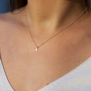 May include: A delicate gold-tone necklace featuring a small, simple cross pendant suspended from a thin, fine link chain. The metal has a polished finish that reflects light, creating a subtle and understated appearance against the white fabric.