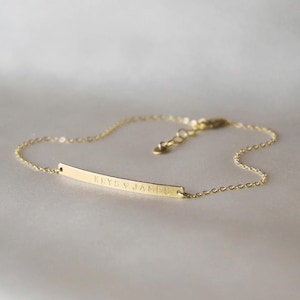Puede incluir: Una delicada pulsera dorada con un colgante de barra curvada y delgada. La barra está grabada con el texto "KRYS & JAMES". La pulsera tiene una fina cadena y un cierre ajustable.