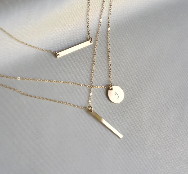 Puede incluir: Tres collares de oro con diferentes colgantes. Un collar tiene un peque&ntilde;o colgante redondo con la letra "J" grabada en &eacute;l. Los otros dos collares tienen colgantes de barra rectangular delgada.