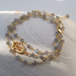May include: A gold chain necklace with a hamsa hand charm and grey labradorite beads.