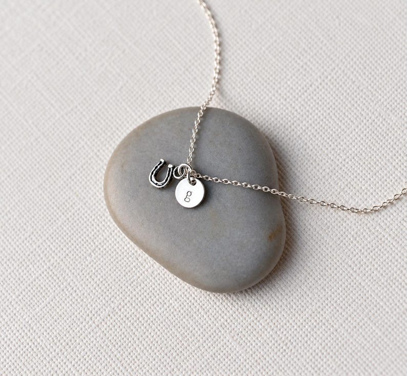 Puede incluir: Un delicado collar de cadena en tono plateado con dos peque&ntilde;os dijes: un colgante de herradura en miniatura y un disco circular grabado con la letra min&uacute;scula g. El collar est&aacute; hecho de finos eslabones met&aacute;licos.