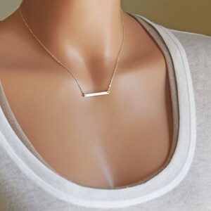 May include: A delicate gold bar necklace with a simple, minimalist design. The necklace is worn by a model with a light brown complexion.