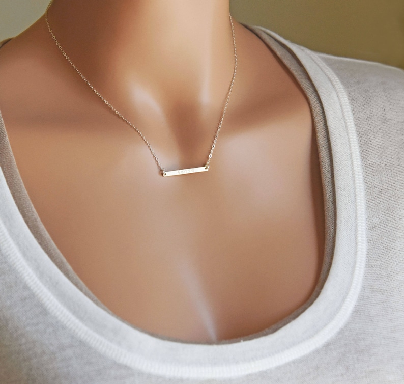 May include: A delicate gold bar necklace with a simple, minimalist design. The necklace is worn by a model with a light brown complexion.