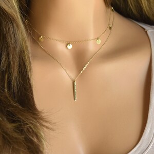 May include: A gold layered necklace with a delicate chain and small round charms. The bottom layer features a longer chain with a vertical bar pendant.