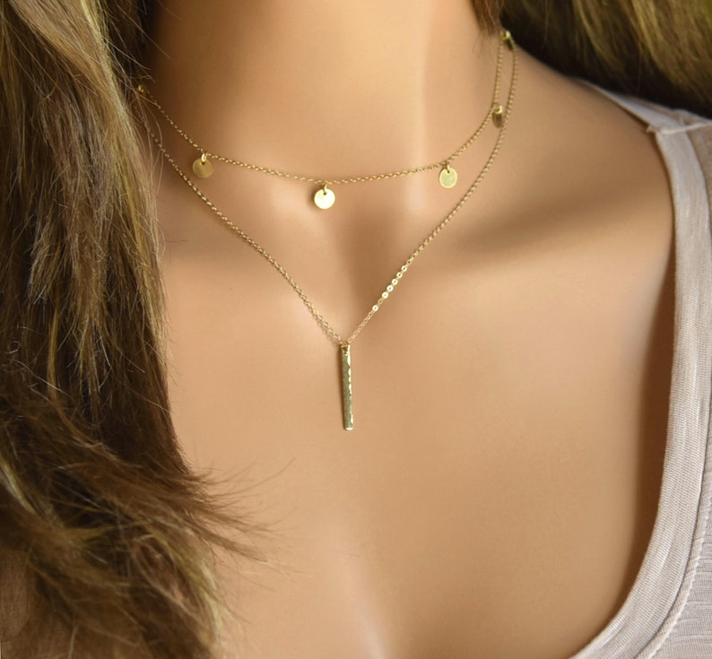 May include: A gold layered necklace with a delicate chain and small round charms. The bottom layer features a longer chain with a vertical bar pendant.