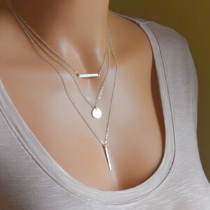 Puede incluir: Un collar de plata de varios niveles con un colgante de barra, un colgante redondo y un colgante largo y delgado.