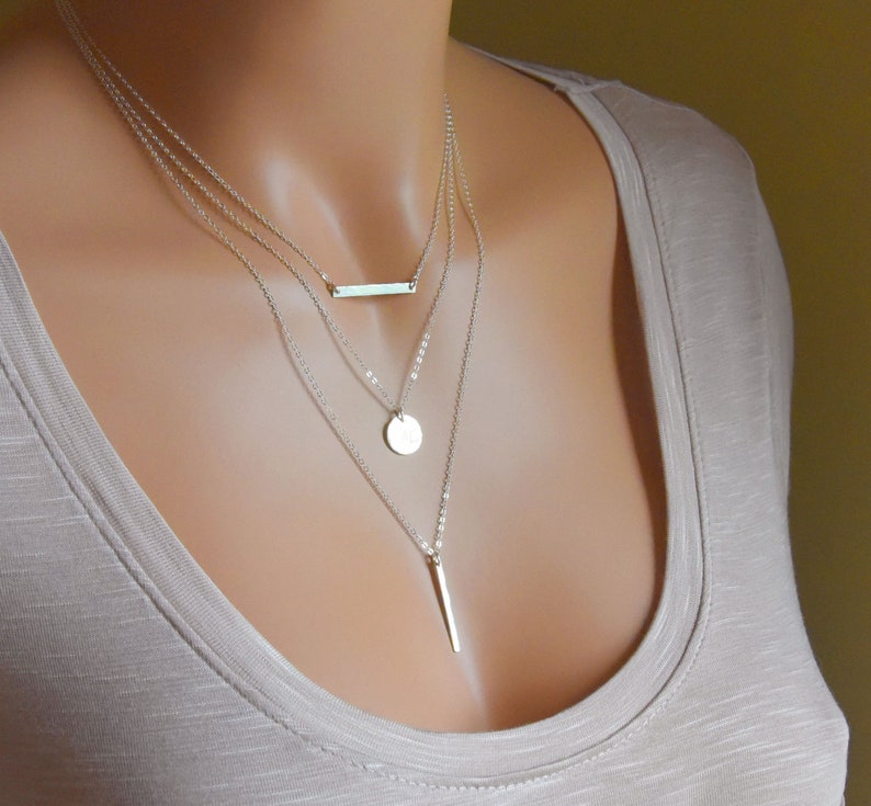 Puede incluir: Un collar de plata de varios niveles con un colgante de barra, un colgante redondo y un colgante largo y delgado.