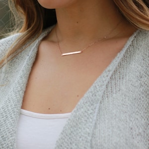 May include: A delicate rose gold necklace with a thin bar pendant. The necklace is worn by a person with long brown hair.