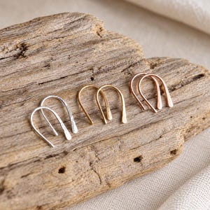 May include: Three pairs of U-shaped earrings in silver, gold, and rose gold tones. Each earring features a hammered, textured finish and a curved, open-ended design. The earrings are arranged in a row, showcasing the distinct metallic colors.