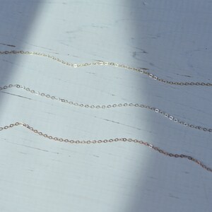 May include: Three delicate gold chains of varying lengths, displayed against a white wooden background.