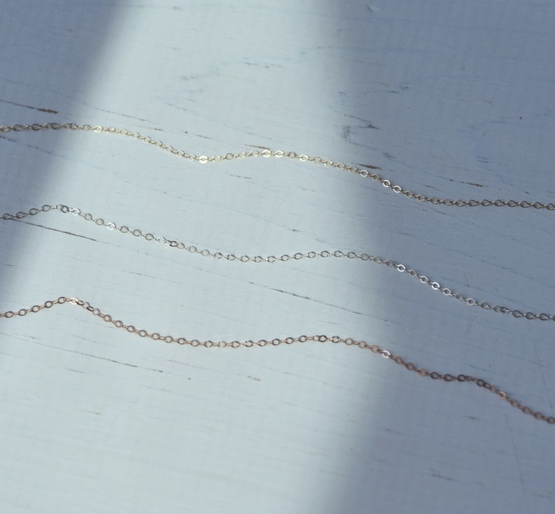 May include: Three delicate gold chains of varying lengths, displayed against a white wooden background.