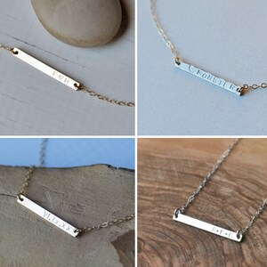 May include: Four gold bar necklaces with different engraved text. The necklaces are on a white and wood background. The text on the necklaces reads "F&H", "&hearts;MONSTIR", "VI.IX.XX", and "E.D.C".