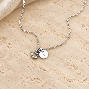 Puede incluir: Un collar de tono plateado con una cadena fina y dos peque&ntilde;os dijes: un dije de herradura con detalles oscuros en relieve y un dije circular grabado con la letra may&uacute;scula V en una tipograf&iacute;a sencilla.