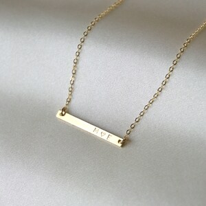 May include: A gold necklace with a thin bar pendant engraved with the letters "M&hearts;E".