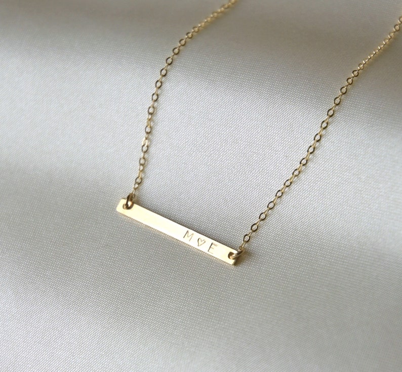 May include: A gold necklace with a thin bar pendant engraved with the letters "M&hearts;E".