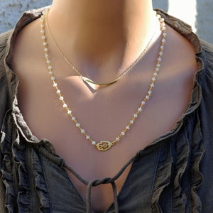 May include: A gold necklace with a hamsa hand charm pendant and a delicate chain. The necklace is layered with a thin gold bar chain.