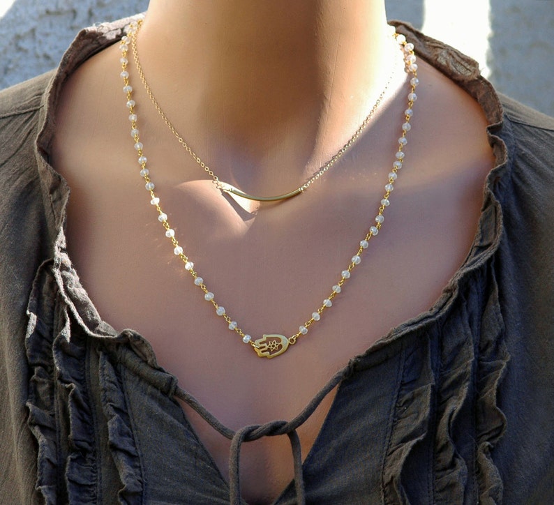 May include: A gold necklace with a hamsa hand charm pendant and a delicate chain. The necklace is layered with a thin gold bar chain.