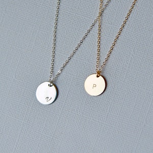 Puede incluir: Dos delicados collares con colgantes redondos. El colgante del collar de la izquierda es de plata y tiene la letra "V" grabada. El colgante del collar de la derecha es de oro y tiene la letra "P" grabada.