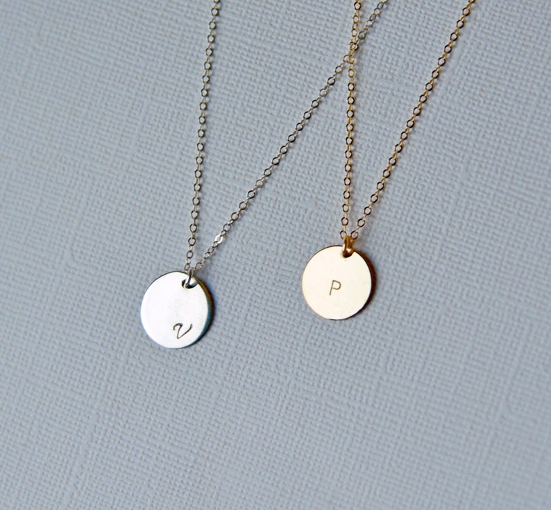 Puede incluir: Dos delicados collares con colgantes redondos. El colgante del collar de la izquierda es de plata y tiene la letra "V" grabada. El colgante del collar de la derecha es de oro y tiene la letra "P" grabada.