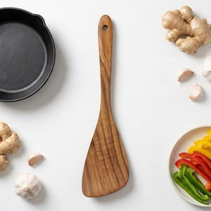 May include: A wooden spatula with a long handle and a wide, flat head is centered between a black cast iron skillet, fresh ginger roots, whole garlic cloves, and a small plate containing sliced red, yellow, and green bell peppers.