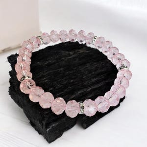 Op de afbeelding: Een roze kralenarmband met facetkralen en zilverkleurige accenten. De armband wordt getoond op een donker, getextureerd oppervlak, dat het delicate ontwerp en de kleur benadrukt. Dit sieraad is een stijlvol accessoire.