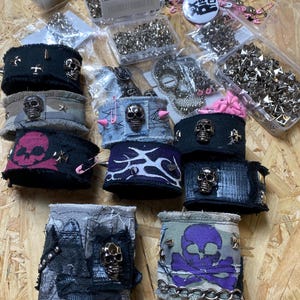 May include: A selection of punk-style wrist cuffs, handcrafted with a variety of materials. The cuffs feature metal studs, skull motifs, and cross designs. Some have pink safety pins and spikes. The colour scheme includes black, grey, and purple. The cuffs are made of denim and canvas.
