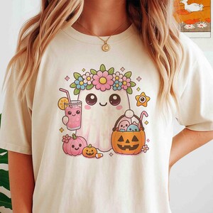 May include: Cream-colored t-shirt featuring a cute ghost character with a floral crown, holding a pink drink with a straw. A jack-o'-lantern basket and pink pumpkins are also in the design. The graphic has a cartoon style with pastel colors.
