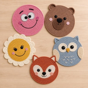May include: Five circular crochet coasters featuring animal and floral designs. The set includes a pink smiley face, a brown bear, a yellow flower with a smiling face, a blue owl, and an orange fox, all crafted with detailed yarn stitching.
