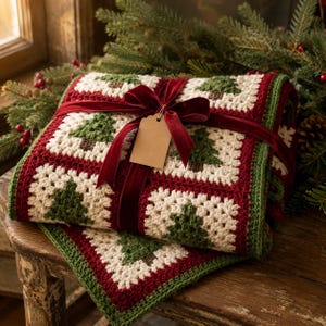 May include: A crocheted blanket featuring a repeating granny square pattern with cream centres and dark green Christmas tree motifs, bordered by deep red and green yarn. The blanket is tied with a dark red velvet ribbon and a blank beige tag.
