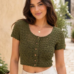 May include: An olive green short-sleeved crochet top featuring a scalloped neckline and hem. The garment has a button-down front with five small brown buttons and a textured, open-work pattern throughout the fabric, ending at the waistline.