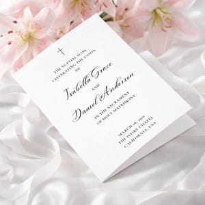 Puede incluir: Un programa de boda blanco con una escritura elegante que anuncia la misa nupcial de Isabella Grace y Daniel Anderson. El programa incluye la fecha, el lugar y un símbolo de cruz. Lirios rosas suaves y tela blanca en el fondo.
