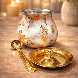 Könnte beinhalten: Ein silberfarbener Metallbehälter mit einem dekorativen Muster aus orangefarbenen und gelben Herbstblättern auf kahlen Zweigen. Das Set enthält einen passenden runden Deckel mit einem zentralen Knauf und einen goldfarbenen Löffel.