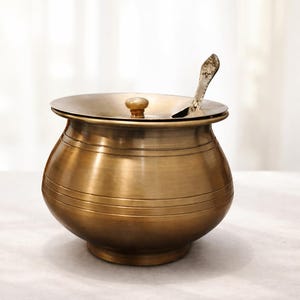 May include: A round brass pot with a smooth, metallic finish and horizontal engraved lines around the body. It features a matching lid with a central knob and a small opening for a decorative metal spoon that rests inside the container.
