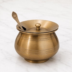 May include: A round brass sugar bowl with a matching lid and a small knob handle. The bowl features horizontal engraved lines around its body and includes a decorative metal spoon with an ornate floral pattern on the handle resting inside.