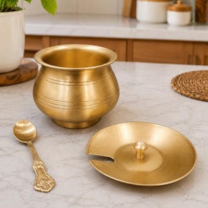 May include: A brass pot with a rounded body and a wide rim, accompanied by a matching circular lid featuring a central knob and a small side notch, alongside a small brass spoon with an ornate handle design and a smooth, polished finish.