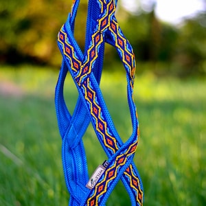 Pode incluir: Uma coleira azul para animais de estimação com um padrão geométrico em amarelo, laranja e azul. A coleira é feita de material tecido e apresenta a marca "WILLGO". O fundo é folhagem verde desfocada.