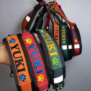 Pode incluir: Várias coleiras para cães em cores vibrantes, incluindo laranja, vermelho e verde. Cada coleira apresenta o nome "YUKI" bordado em uma cor contrastante, juntamente com uma pegada de pata. As coleiras têm faixas refletivas.