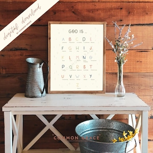 Puede incluir: Una impresión enmarcada con el texto "GOD IS..." y un alfabeto con palabras como "Awesome" y "Believe". La impresión está sobre una mesa blanca con una jarra de metal y un cubo galvanizado. El fondo es una pared de madera.