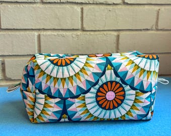 May include: A rectangular cosmetic bag with a geometric pattern in teal, orange, pink, and yellow. The bag has a zip closure and small silver pulls. The background is a light blue surface and a white brick wall.