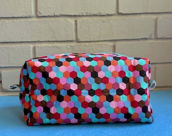 May include: A rectangular make-up bag with a vibrant geometric pattern of hexagons in red, orange, blue, pink, and brown. It features a zip closure and small fabric loops on each end.