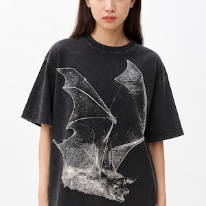 May include: Dark grey t-shirt with a large, detailed white bat graphic. The bat's wings are spread wide. The shirt has a faded, vintage look. The design is centrally placed, making it a bold statement piece.