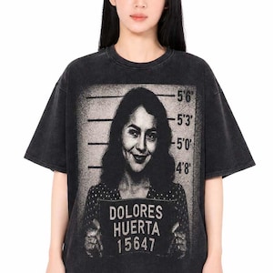 May include: Black t-shirt featuring a grayscale image of Dolores Huerta holding a sign with her name and the number 15647. The shirt has a vintage, washed look.