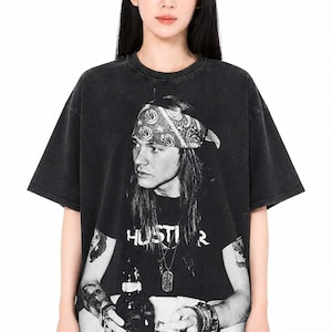 Op de afbeelding: Zwart-wit grafisch T-shirt met een portret van een man met een bandana, met het woord "HUSTLER" eronder gedrukt. Het shirt heeft korte mouwen en een ronde hals.