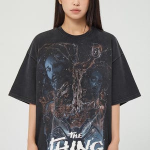 May include: Black t-shirt with a graphic from the film "The Thing." The design features characters and the film's title in a blue and white font. The shirt has a vintage, washed appearance.