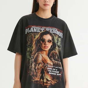 May include: Black Planet Terror t-shirt with a movie poster design. The graphic features a woman in sunglasses, the film title, and the text "Humanity's last hope rests on a high power machine gun." The shirt has a vintage wash.