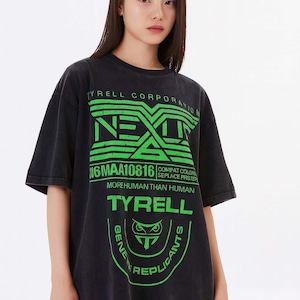 Puede incluir: Camiseta negra con un diseño gráfico verde. El diseño incluye el texto "TYRELL CORPORATION", "NEXT", "N6MAA10816", "MORE HUMAN THAN HUMAN" y "TYRELL". La camiseta tiene un aspecto vintage y desgastado.