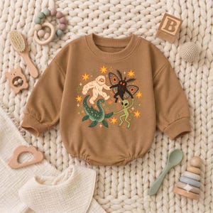 May include: A tan long-sleeve baby romper featuring a central graphic print of a white yeti, a black moth-like creature, a green sea monster, and a green alien holding hands, surrounded by orange stars and small green foliage elements.